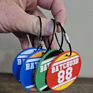 May include: Four circular plastic luggage tags in blue, orange, green, and red, each featuring white raised text that reads BATCHO3D and the number 88. Each tag is attached to a black flexible cable loop with a silver metal screw fastener.