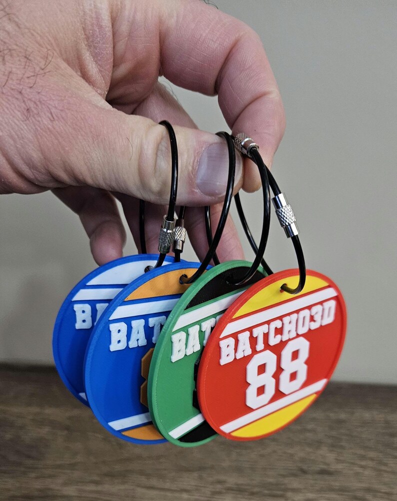 May include: Four circular plastic luggage tags in blue, orange, green, and red, each featuring white raised text that reads BATCHO3D and the number 88. Each tag is attached to a black flexible cable loop with a silver metal screw fastener.