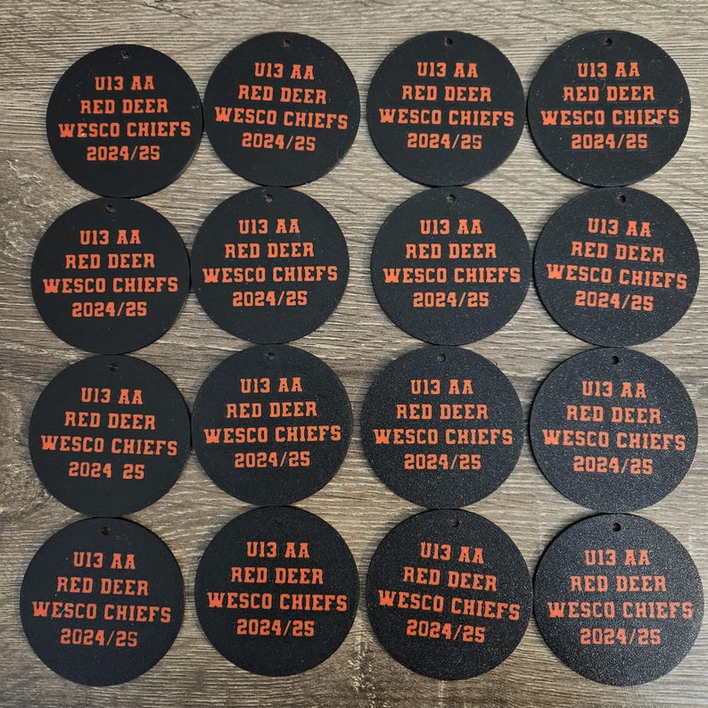 May include: Sixteen circular black ornaments featuring orange text that reads U13 AA RED DEER WESCO CHIEFS 2024/25. Each ornament has a small hole at the top for hanging. The design is consistent across all sixteen individual round pieces.