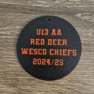 May include: A circular black ornament featuring orange text that reads U13 AA RED DEER WESCO CHIEFS 2024/25. The object has a small hole at the top for hanging and a textured surface finish, displaying team information in a bold, block-style font.