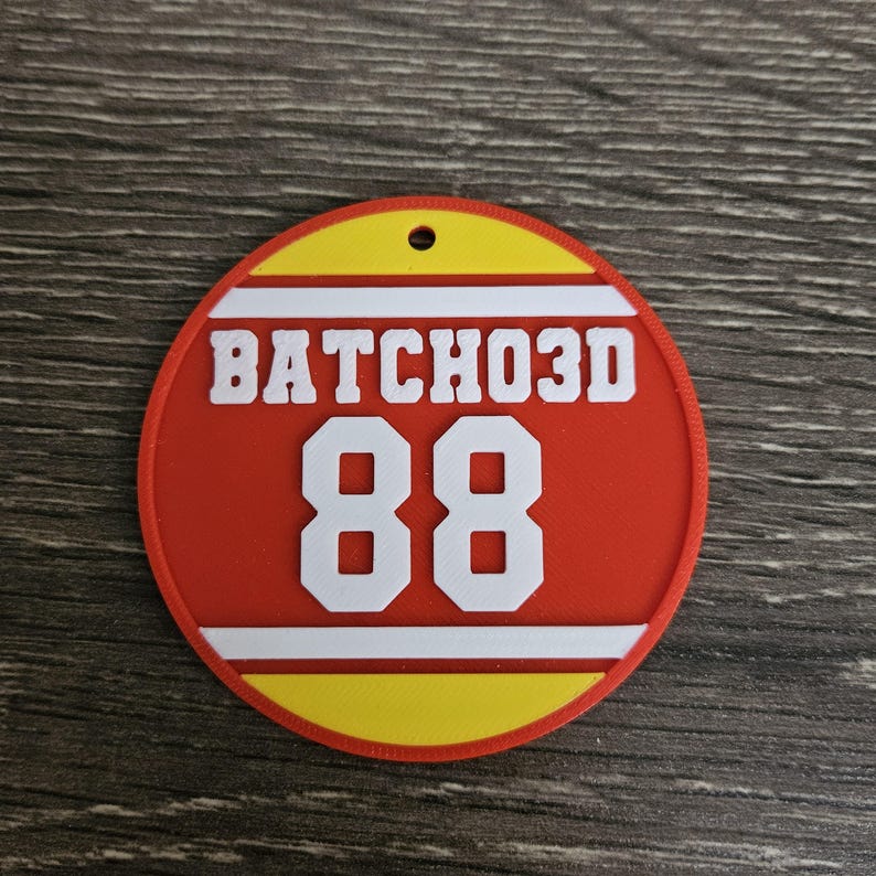 May include: A circular red, yellow, and white plastic tag featuring the text BATCHO3D in white lettering above the large white number 88. The tag has a small hole at the top center for hanging and a textured surface finish throughout.