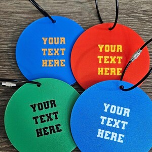 May include: Four circular luggage tags in blue, red, green, and light blue, each featuring a black metal cable loop. The tags are embossed with the text YOUR TEXT HERE in contrasting colors, including yellow, black, and white lettering.