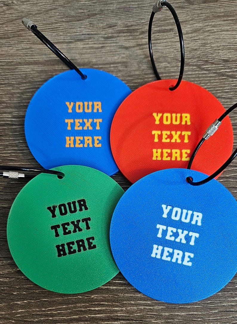 May include: Four circular luggage tags in blue, red, green, and light blue, each featuring a black metal cable loop. The tags are embossed with the text YOUR TEXT HERE in contrasting colors, including yellow, black, and white lettering.