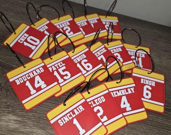 May include: A collection of rectangular red and yellow luggage tags, each featuring a white sports jersey-style design with a surname and a number. Each tag is attached to a black metal ring, displaying various names like Chen, Patel, and Singh.