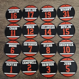 May include: Sixteen circular black ornaments featuring white horizontal stripes, orange numbers, and white surnames including Martin, Darling, Johnson, Wright, Craig, Birch, Kelahear, Devlin, Bosse, Van Grinsven, Stang, Sherwick, Rentas, Campbell, Dawson, and Jubb.