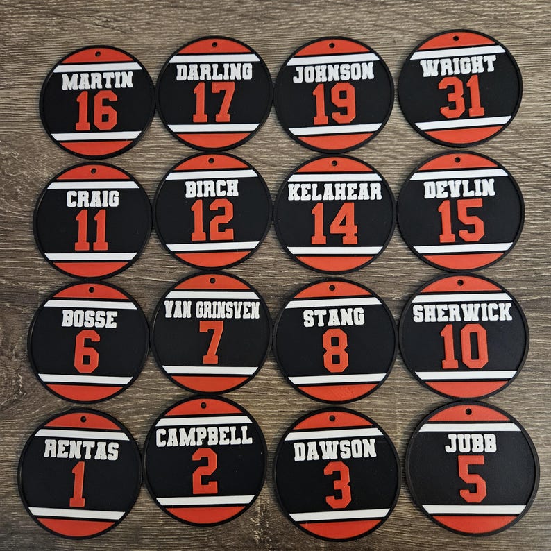 May include: Sixteen circular black ornaments featuring white horizontal stripes, orange numbers, and white surnames including Martin, Darling, Johnson, Wright, Craig, Birch, Kelahear, Devlin, Bosse, Van Grinsven, Stang, Sherwick, Rentas, Campbell, Dawson, and Jubb.