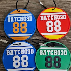 May include: Four circular luggage tags with black wire loops. Each tag features the text BATCHO3D and the number 88. The tags are blue with orange, red with yellow, blue with white, and green with black, all displaying white or black accents.