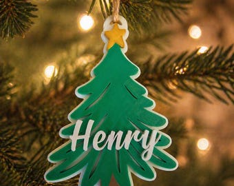May include: A green Christmas tree ornament with the name "Henry" in white script. The ornament has a yellow star at the top and a brown base. It is hanging from a natural twine string, perfect for holiday decor.