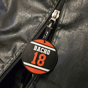 May include: A circular black and orange rubber zipper pull attached to a black textured bag. The tag features the white text BACHO above the large orange number 18, framed by horizontal white stripes on a black background with orange borders.