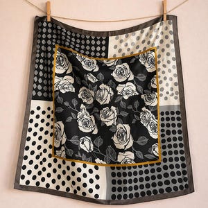 May include: Square silk scarf with a black and white rose pattern in the center, framed by a gold border. The corners feature polka dots in black, white, and gray, with a dark gray border. The scarf is hanging from a clothesline.