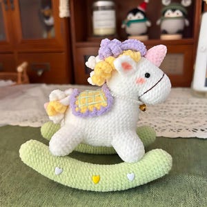 May include: A handmade white crochet unicorn rocking horse with a light green base. The unicorn has a purple mane, yellow accents, and a small gold bell. The rocking base has heart-shaped decorations.
