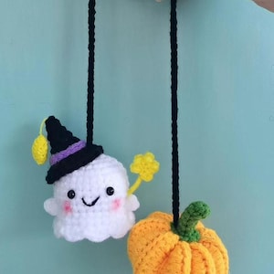 May include: Crocheted Halloween decorations featuring a white ghost wearing a witch hat and holding a yellow star wand, and an orange pumpkin. The ghost has rosy cheeks and a smiling face. The decorations are suspended by black yarn.