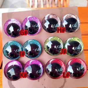 May include: A set of eight round, domed craft eyes arranged in pairs on a brown card. The eyes feature large black pupils surrounded by glittery irises in purple, silver, teal, green, and red. Each pair is labeled with letters G#, H#, I#, J#, K#, and L#.