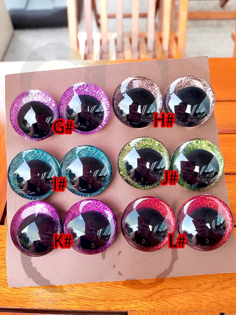 May include: A set of eight round, domed craft eyes arranged in pairs on a brown card. The eyes feature large black pupils surrounded by glittery irises in purple, silver, teal, green, and red. Each pair is labeled with letters G#, H#, I#, J#, K#, and L#.