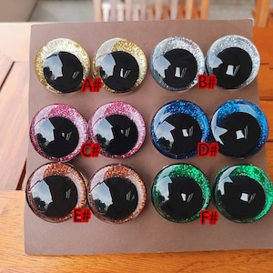 May include: Twelve circular craft eyes arranged in pairs on a brown card, labeled A# through F#. Each eye features a large, glossy black pupil surrounded by a vibrant, glittery iris in gold, silver, pink, blue, copper, or green color variations.