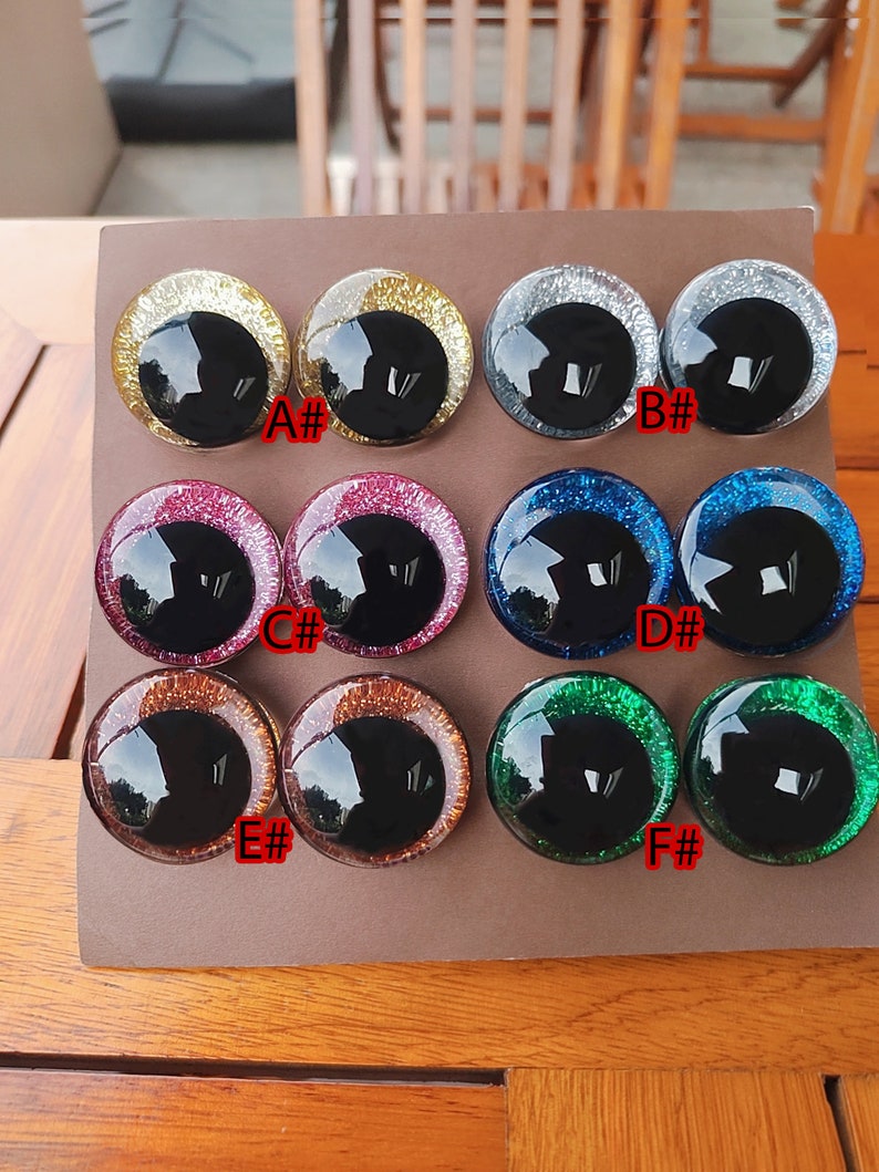 May include: Twelve circular craft eyes arranged in pairs on a brown card, labeled A# through F#. Each eye features a large, glossy black pupil surrounded by a vibrant, glittery iris in gold, silver, pink, blue, copper, or green color variations.