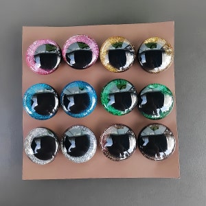 May include: Twelve circular cabochons arranged in a grid, each featuring a smooth, black, reflective center surrounded by a wide, glittery border. The colors of the glitter borders include pink, gold, blue, green, silver, and bronze tones.