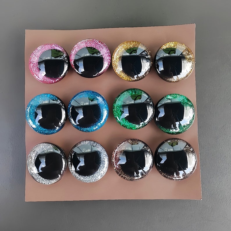 May include: Twelve circular cabochons arranged in a grid, each featuring a smooth, black, reflective center surrounded by a wide, glittery border. The colors of the glitter borders include pink, gold, blue, green, silver, and bronze tones.