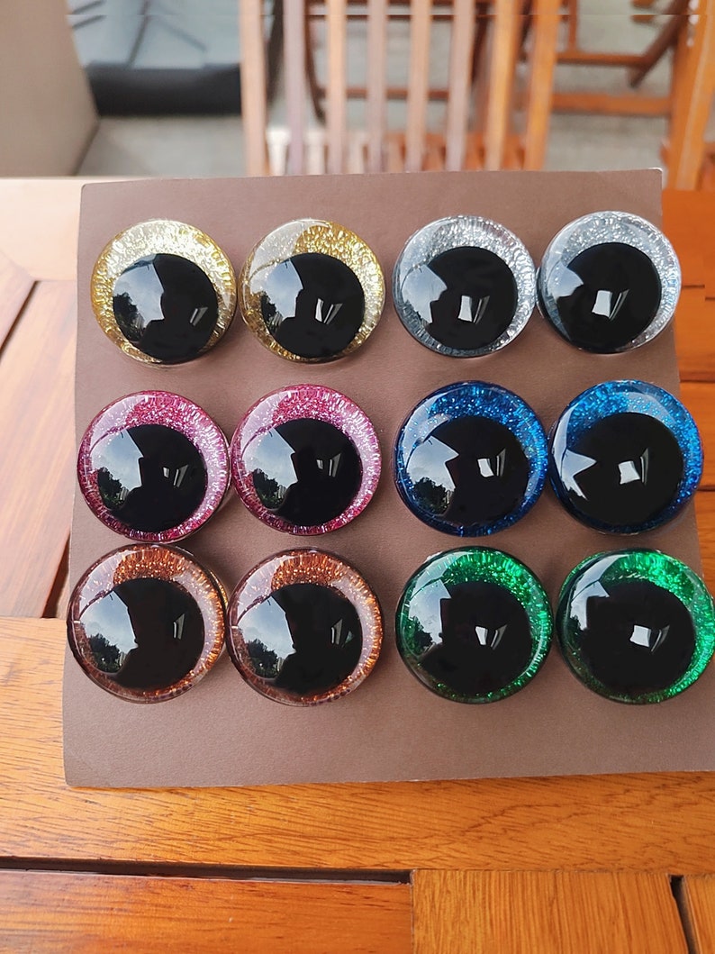 May include: A set of twelve circular craft eyes arranged in three rows of four on a brown display card. Each eye features a large, glossy black pupil surrounded by a vibrant, glittery iris in gold, silver, pink, blue, copper, and green colors.