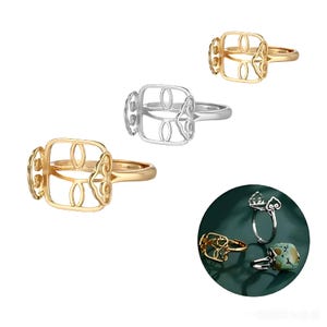 May include: A collection of open-design rings in gold and silver tones. The rings feature a square or rectangular setting with decorative cutouts. One ring has a turquoise stone. The rings are displayed on a white background.
