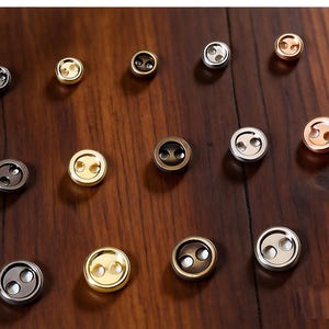 May include: A collection of small round metal buttons arranged in rows, labelled 3mm, 4mm, and 5mm. The buttons feature two holes and come in various metallic finishes including silver, gold, bronze, and copper tones, displayed on a dark wood surface.