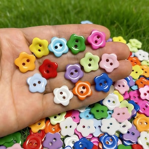 May include: A collection of small, flower-shaped buttons in a variety of bright colors including red, orange, yellow, green, blue, purple, pink, and white. Each button features two holes in the center for sewing and a smooth, glossy finish.