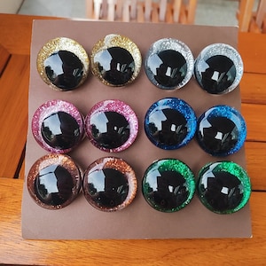 May include: A set of twelve circular craft eyes arranged in three rows of four on a brown display card. Each eye features a large, glossy black pupil surrounded by a vibrant, glitter-infused iris in gold, silver, pink, blue, copper, and green.