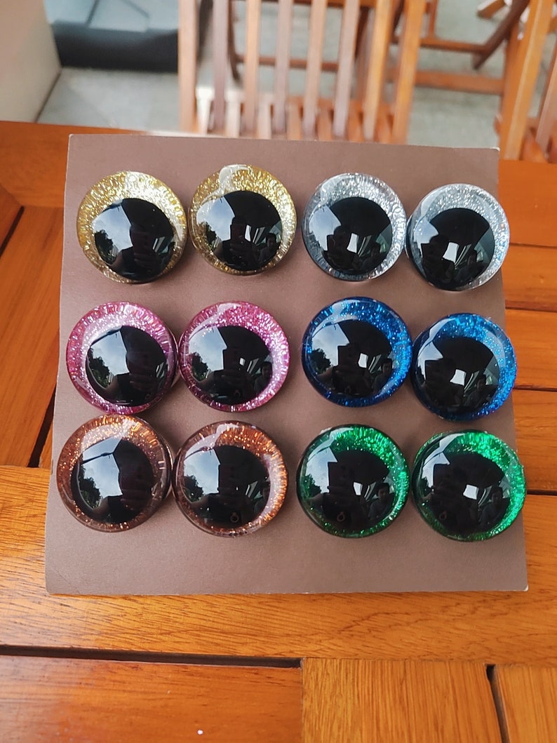 May include: A set of twelve circular craft eyes arranged in three rows of four on a brown display card. Each eye features a large, glossy black pupil surrounded by a vibrant, glitter-infused iris in gold, silver, pink, blue, copper, and green.