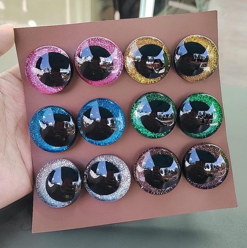 May include: A collection of twelve circular cabochons arranged in three rows on a brown card. Each piece features a large black pupil surrounded by a vibrant, glittery iris in colors including pink, gold, blue, green, silver, and purple.