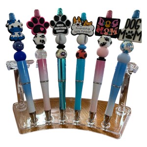 May include: Six colorful pens with different charms on top, including a paw print, a dog, a cow, and a heart. The pens are displayed on a clear acrylic stand.