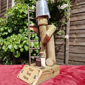 May include: A handcrafted wooden figure of a painter, wearing a silver bucket as a hat, with a ladder and a small bucket. The base has a sign that reads "Your Text Here".