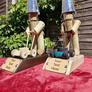 May include: Two wooden lawn mower figurines wearing bucket hats. One has a wooden mower, the other a black and teal mower. Each figurine has a sign that says "Your Text Here". The figurines are on a red surface.