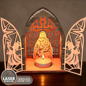 May include: A laser-cut wooden religious scene featuring a mother and child, with a cross design and angel cutouts. A lit candle sits in front of the figures. The design includes the text "LASER BUILD.LAB TEMPLATE LASER CUT".