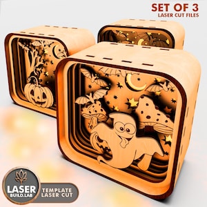 Könnte beinhalten: Set aus drei lasergeschnittenen Halloween-Schattenboxen aus Holz. Jede Box hat geschichtete Designs mit Kürbissen, Geistern, Fledermäusen, Pilzen, Sternen und Monden. Die Boxen sind aus hellem Holz mit warmem orangefarbenem Glanz gefertigt.