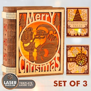 Pode incluir: Um conjunto de três decorações de Natal em madeira. A peça principal é um design em forma de livro com um Pai Natal cortado a laser e as palavras "Merry Christmas". Duas peças menores apresentam cenas de Natal e os textos "Christmas Tales" e "XMAS".