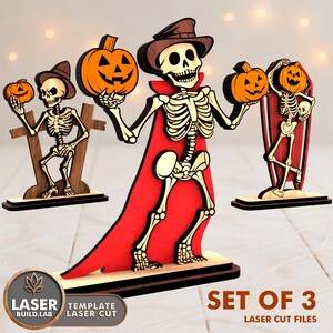 May include: Set of 3 laser-cut Halloween decorations featuring skeleton figures holding jack-o'-lanterns. The skeletons are wearing hats and capes, and are made of wood with a red and orange color scheme. The text "SET OF 3" is visible.