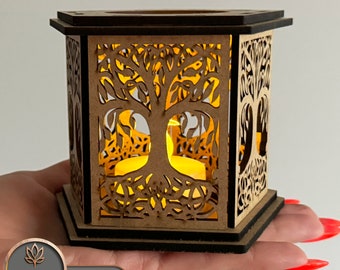 May include: A wooden candle holder featuring a laser-cut tree of life design. The holder is hexagonal in shape and has a light brown finish. A tea light candle is lit inside the holder.