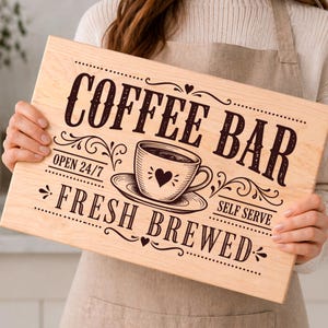 Può includere: Un cartello in legno chiaro con le parole "COFFEE BAR" in lettere marroni scure. Il cartello presenta una grafica di tazza di caffè con un cuore e le frasi "OPEN 24/7", "FRESH BREWED" e "SELF SERVE".