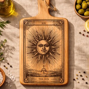 Puede incluir: Tabla de cortar de madera con un diseño de carta del tarot que representa el sol. La tabla está rodeada de ingredientes como aceitunas, limones, hierbas y especias. La carta del sol tiene el texto "XIX" y "THE SUN".