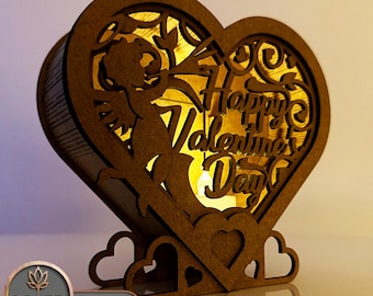 May include: A wooden heart-shaped box with a cutout design of a cupid holding a heart and the text "Happy Valentine's Day". The box has a light inside and is illuminated with a warm glow.