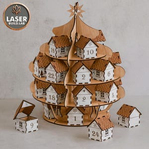 Op de afbeelding: Houten adventskalender in de vorm van een kerstboom. Het heeft kleine huisjes met nummers van 1 tot 24. De huisjes hebben bruine daken en witte muren. De boom is bekroond met een ster. Het logo "LASER BUILD.LAB" is zichtbaar.