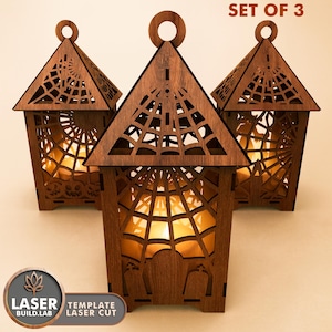 May include: Three wooden Halloween lanterns with intricate laser-cut designs. Each lantern features a triangular roof with a spiderweb pattern, a circular handle, and a warm, glowing light inside. The text "SET OF 3" is visible.