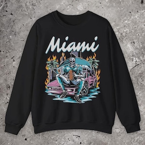 Puede incluir: Sudadera negra con un gráfico de un jugador de fútbol americano esqueleto sentado en un coche rosa. La palabra "Miami" está escrita en blanco sobre el gráfico. Hay palmeras y llamas en el fondo.