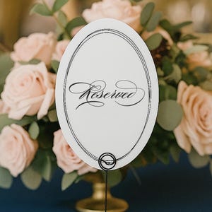 May include: An oval white table sign featuring a black double-line border and the word Reserved written in elegant black script. The sign is held upright by a thin black metal wire stand with a circular base, positioned on a dark blue surface.