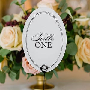 May include: A wedding table setting with a floral centerpiece and a table number. The white oval table number reads "Table ONE" in elegant script. A folded napkin and plate are in the foreground.