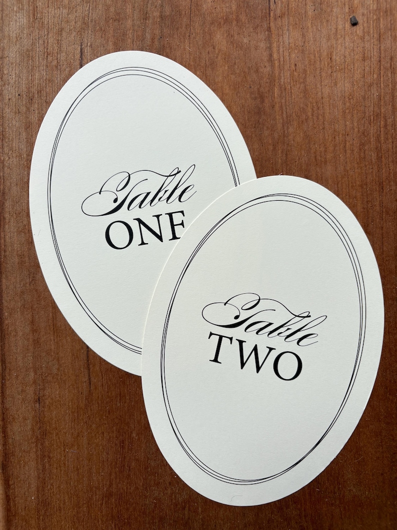 Op de afbeelding: Twee ovale tafelkaarten met zwarte tekst en een dubbele zwarte rand. E&eacute;n kaart leest "Table ONE" en de andere "Table TWO" in elegant schrift. De kaarten liggen op een houten oppervlak.
