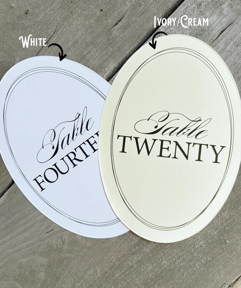 Op de afbeelding: Twee ovale tafelnummerkaarten, &eacute;&eacute;n witte en &eacute;&eacute;n ivoor/cr&egrave;me. De witte kaart leest "Table FOURTEEN" in elegant zwart schrift. De ivoorkleurige kaart leest "Table TWENTY" in hetzelfde schrift. Beide kaarten hebben een dunne zwarte rand.