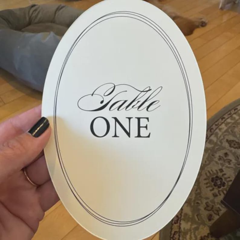 Op de afbeelding: Een ovaalvormige, cr&egrave;mekleurige tafelnummerkaart met de woorden "Table ONE" in elegant zwart schrift. De kaart is omrand door een dunne, dubbele zwarte lijn. Een hand met zwarte nagellak houdt de kaart vast.
