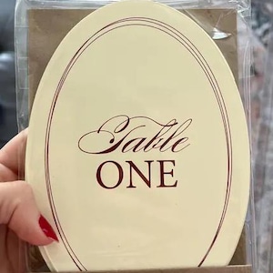 Op de afbeelding: Een ovale tafelkaart met het woord "Table" in elegant schrift en "ONE" in vette letters. De kaart heeft een cr&egrave;mekleurige achtergrond met een dunne, donkerrode rand en tekst. De kaart is verpakt in helder plastic.