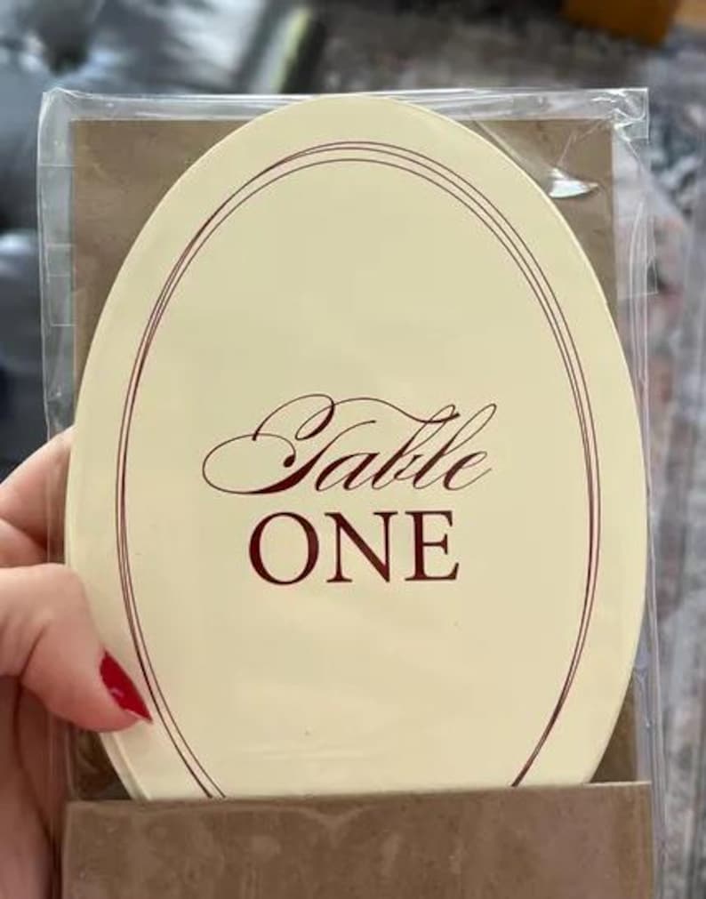 Op de afbeelding: Een ovale tafelkaart met het woord "Table" in elegant schrift en "ONE" in vette letters. De kaart heeft een cr&egrave;mekleurige achtergrond met een dunne, donkerrode rand en tekst. De kaart is verpakt in helder plastic.
